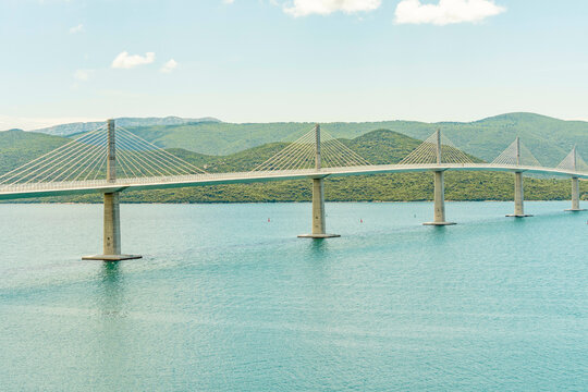 Coastal landscape featuring a long suspension Peljesac Bridge spanning across a blue bay with a lush green shoreline under a clear sky, Peljesac, Croatia