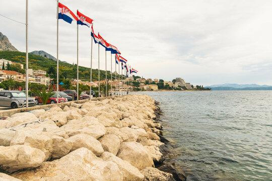 Coastal scene with a row of Croatian flags fluttering on masts beside a stone embankment with parked cars and a cloudy sky backdrop by the Adriatic Sea, Podgora, Croatia