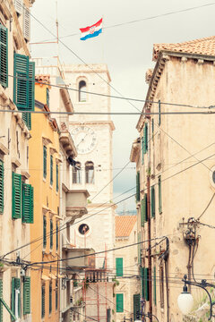 Narrow street with traditional houses and a clock tower flying the Croatian flag under a cloudy sky, Sibenik, Croatia