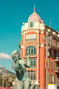 A bronze statue in front of a vibrant orange building with a colorful tiled dome under a clear blue sky by Fontaine du Soleil by Place Massena, Nice, France  