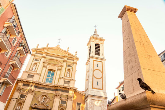 A pigeon perches on a sunlit ledge with a baroque church and an obelisk in the background against a clear blue sky, Nice, France