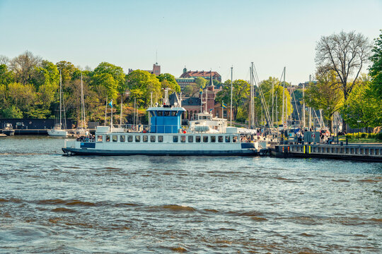 A blue and white ferry named at a pier with people and sailboats in the background on a sunny day by Skeppsholmen, Stockholm, Sweden
