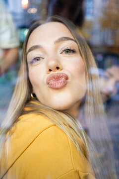 Woman in a yellow top pouting her lips for a kiss behind a glass pane with reflections and blurry background, Berlin, Germany