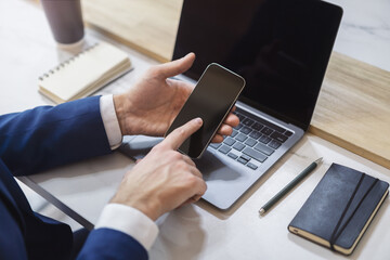 Man engaged with his modern smartphone, laptop slightly out of focus nearby against the blurred...