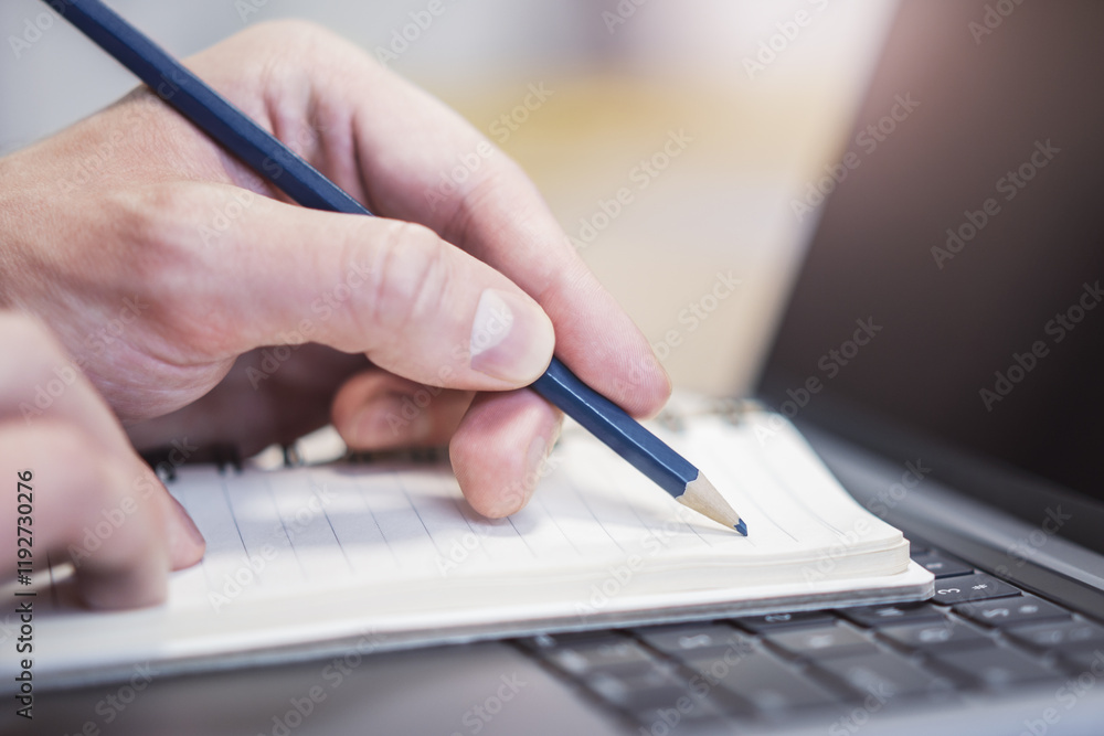 Aufkleber focused view of a man's hand as he writes in a notepad on a modern laptop, background gently blurred