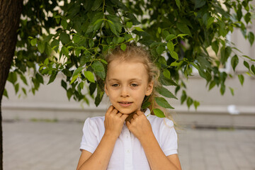 Thoughtful young girl, wearing a white shirt, standing under a tree with green leaves. She is...