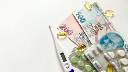 A conceptual arrangement of Turkish lira banknotes (200 and 100 denominations), a digital thermometer, and various pills and capsules scattered across a white background. 