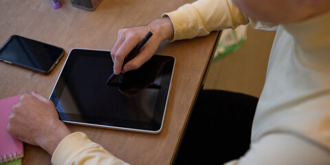 Obraz premium Person sitting at desk, holding stylus drawing on tablet. Workspace featuring notebooks, pens, and a smartphone. Digital art and education. Concept of business, modern technologies, gadgets