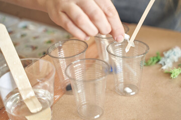 Hands preparing resin mixture with wooden sticks in clear plastic cups on a crafting table.