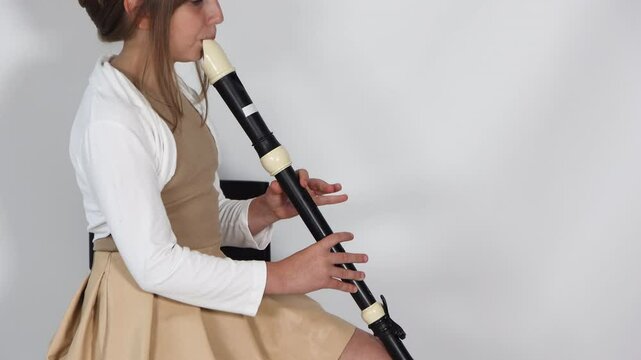 Girl playing, practicing the flute, playing a melody on a musical instrument, wooden pipe, sitting on black chair on white background. Out-of-school children activities, lessons