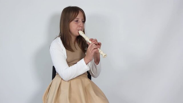 Girl playing, practicing the flute in bright room sitting on black chair on white background. Children musicians concept. Out-of-school activities, home distance learning.