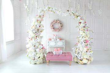 White living room interior with pink sofa pouf and decorative blossom flowers arch. Holiday spring decor. Easter festive decoration with wreath on wall, chest of drawers, ceramic rabbit and easter egg