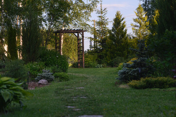Beautiful summer garden view with curvy stone pathway and wooden archway (pergola). Natural rustic cottage garden with hostas, conifers and shrubs