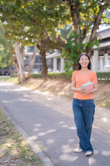 Fototapeta premium Happy Asian student walking through a university campus, holding books and smiling brightly while gazing off into the distance, enjoying a beautiful day filled with hope and inspiration