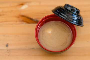 A bowl of miso soup served in a red and black traditional container with a wooden table