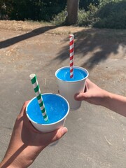 Two hands holding cups of blue frozen slushies with paper straws. 
