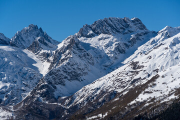 Dombay. Karachay-Cherkessia. Giant rocks with snow-capped peaks and glaciers rise majestically against  blue autumn sky. Pine and spruce forests grow at foot of mountains. Sunny day. Autumn 2024
