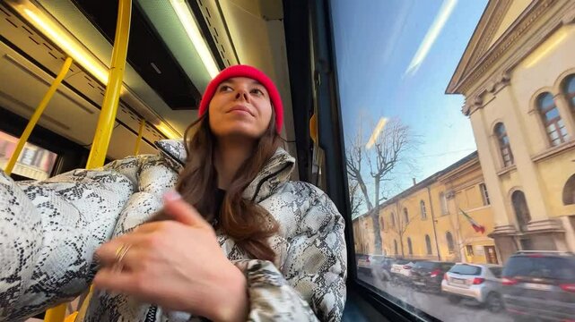 Verona, Italy - 12 01 2025: caucasian brunette girl in red trandy cap and snake print down jacket riding on the bus and looking the window in Italian Verona in sunny day
