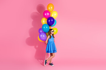 Smiling young girl holding colorful balloons in a blue dress on a pink background, representing happiness and celebration.