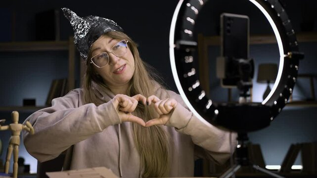 Strange crazy vlogger with cellphone, paranoid woman streaming at home conspiracy theory broadcasting in vlog, showing heart shape gesture with love emotion. blogging and conspiracy theory