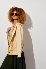 Fashion model with curly hair wearing a cozy cardigan and sunglasses against a plain white background, showcasing a stylish casual look with a playful vibe