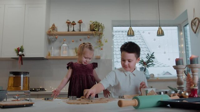 Children take part in making Christmas gingerbread cookies. Boy enthusiastically cuts out dough figures with molds. Little girl discontentedly shakes flour off her hands and dress. Slow motion