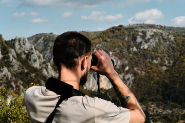 Young hiker man using binoculars while standing on a lookout in autumn mountains. Traveler guy holding binoculars outdoor. A man watches birds and wildlife. Rear view portrait.
