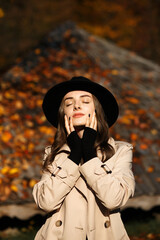 woman with long brown hair. She stands against the background of an autumn landscape, where trees with yellow and orange leaves are visible. The woman is wearing a light raincoat and a black hat.