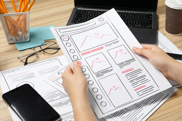 Website designer holding wireframe at wooden table with smartphone and laptop, closeup