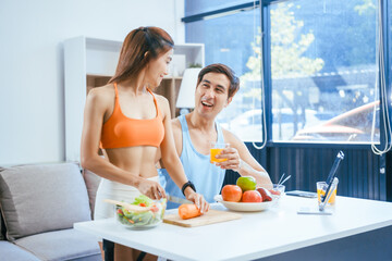 A happy young couple in sportswear sits on the sofa after exercising, enjoying fruits, salad, and orange juice. They focus on weight loss, weight control, and living a healthy lifestyle