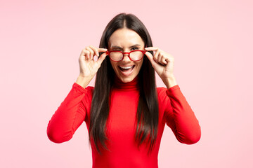 Playful Latin woman winking and adjusting red glasses on pink background