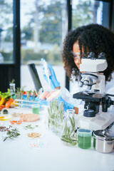 An African American female scientist works at her desk in a laboratory, closely examining a meat sample under a microscope.She focuses on modification,plant genetics,innovations in food meat research