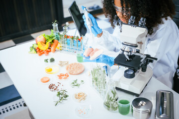 An African American female scientist works at her desk in a laboratory, closely examining a meat sample under a microscope.She focuses on modification,plant genetics,innovations in food meat research