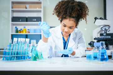 An African American female scientist in a white coat and blue gloves smiles confidently, holding a...