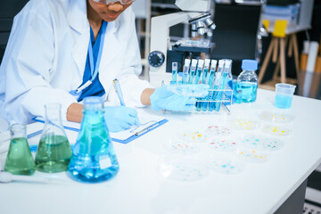 Fototapeta premium An African American female scientist in a white coat and blue gloves smiles confidently, holding a test tube with blue liquid. She conducts drug testing and medical research in a laboratory