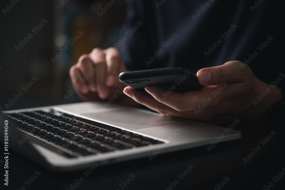Poster man holding a smartphone while typing on a laptop keyboard in an office or home setting, representin