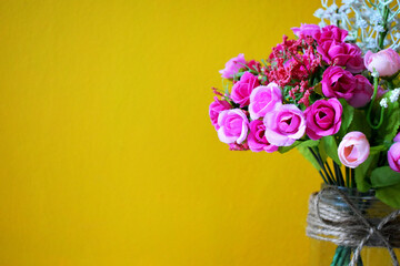A bouquet of pink artificial roses in a glass vase against a yellow background.