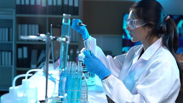 Professional female scientist in white coat and protective glasses carefully adding blue liquid using a pipette to test tubes in modern laboratory with advanced equipment
