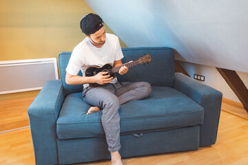 Man with talent sits on a blue sofa, carefully tuning a black ukulele. Musician is enjoying the...