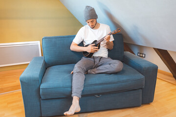 Man with talent sits on a blue sofa, carefully tuning a black ukulele. Musician is enjoying the...