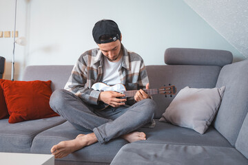 Man with talent sits on a gray sofa, carefully tuning a black ukulele. Musician is enjoying the...