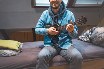 Obraz premium Musician with smile in a denim jacket and hoodie sits on a cozy bed, strumming a black ukulele. Enjoying the feeling of playing the ukulele and immersing oneself in the creative spirit