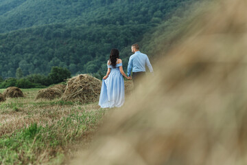 couple walking through a field with hay. The woman is wearing a long blue dress with open shoulders, and the man is wearing a light blue shirt and dark trousers.