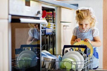 Little adorable cute toddler girl helping to unload dishwasher. Funny happy child standing in the...