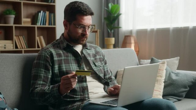 Confused adult man male guy shopping laptop at home couch payment online buying internet purchase holding credit card web money e-commerce using computer problem failure unsuccessful transaction error
