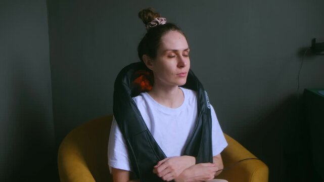 Woman with closed eyes uses warm back massager from muscle tension at home. Female undergoes physical injury recovery with electric device