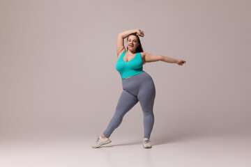 Chubby woman in blue top and leggings raising one arm overhead while smiling, standing in dynamic...