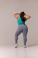 Obraz premium rear view photo of chubby woman in blue top and leggings posing confidently, holding hair with hands against white background. Concept of beauty, fitness, movement, dance and music.