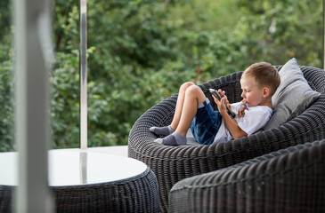 A 5-year-old little boy with a smartphone in his hands on the veranda. The child is playing on the phone. Harm of gadgets for children. Protect your child from the smartphone. Kids and gadgets.