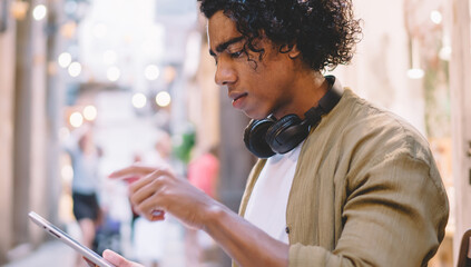 Pensive curly man in casual wear sitting on city street reading news from social networks using digital tablet, serious handsome male installing application on touchpad enjoying online chatting
